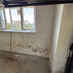 Złota rączka - Wnętrze zaniedbanego pokoju z odpadającą farbą, brudną podłogą i otwartym oknem z widokiem na zabudowę. W rogu stoi miotła.