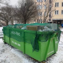 Złota rączka - Zielony kontener Eco-Tec, wypełniony odpadami i zabezpieczony siatką, stoi na śniegu przed blokiem mieszkalnym w pochmurny dzień. Widoczne numery kontaktowe na kontenerze.