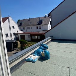 Złota rączka - Błękitne worki i płyty na zielonym dachu płaskim, widok z okna na zabudowę mieszkalną w słoneczny dzień. Prace porządkowe na dachu.