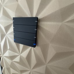AUTOMATYK SP. Z O.O - Czarny panel sterowania inteligentnego domu z subtelnym, niebieskim podświetleniem LED, zamontowany na dekoracyjnej ścianie z trójwymiarową fakturą.