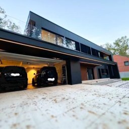 AUTOMATYK SP. Z O.O - Nowoczesny dom z garażem, oświetleniem LED i dwoma samochodami. Widok z dołu, podkreślający architekturę i oświetlenie zewnętrzne.