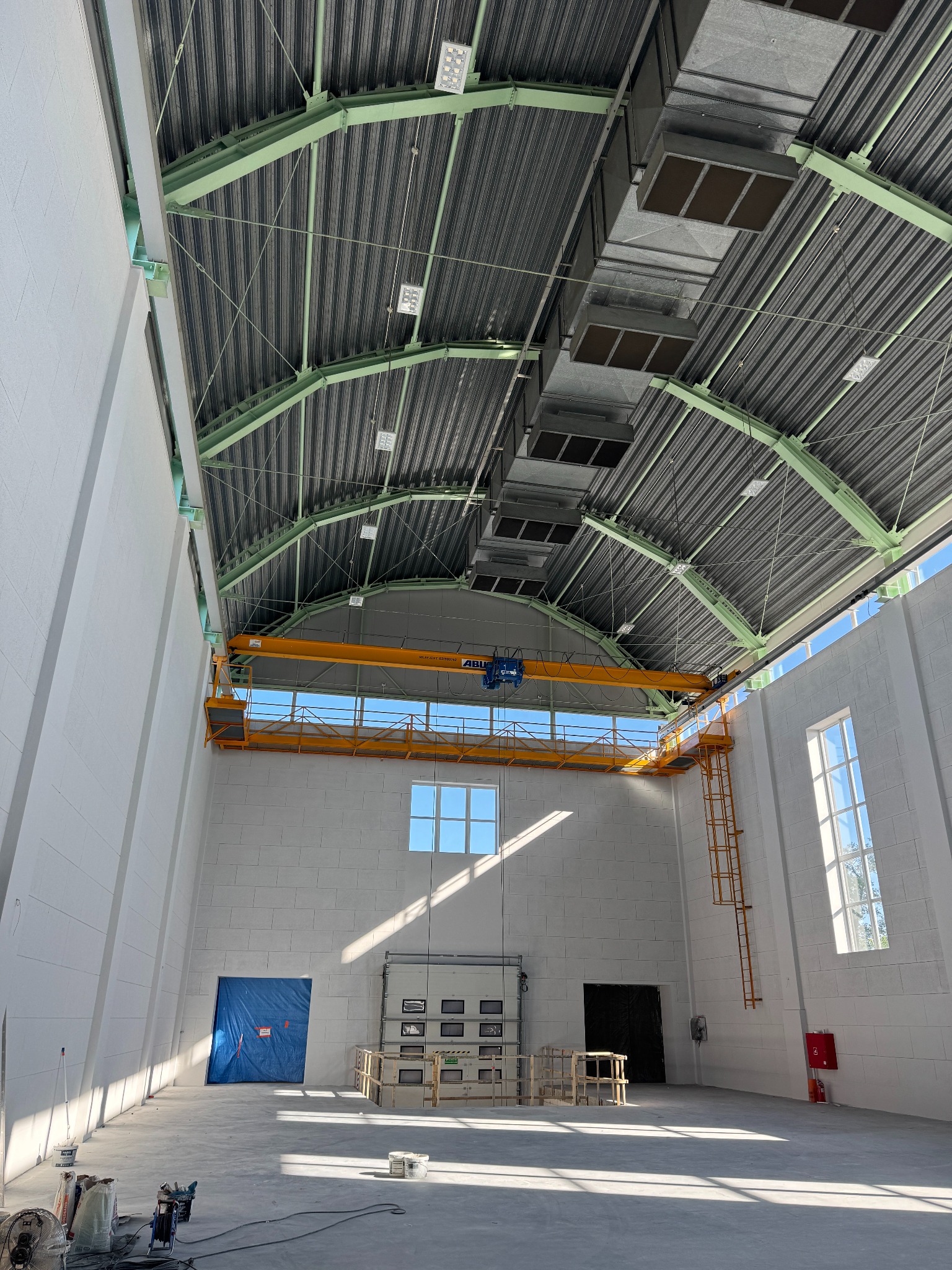 Wnętrze hali przemysłowej z suwnicą, widoczne instalacje pod stropem, okna i drzwi. Ściany białe, podłoga betonowa. Widoczne elementy instalacji elektrycznych.