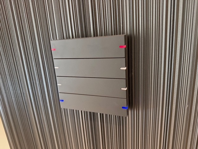 Panel sterowania inteligentnego domu, cztery poziome segmenty z kolorowymi znacznikami (czerwony, biały, niebieski) na tle ściany z pionowymi paskami.