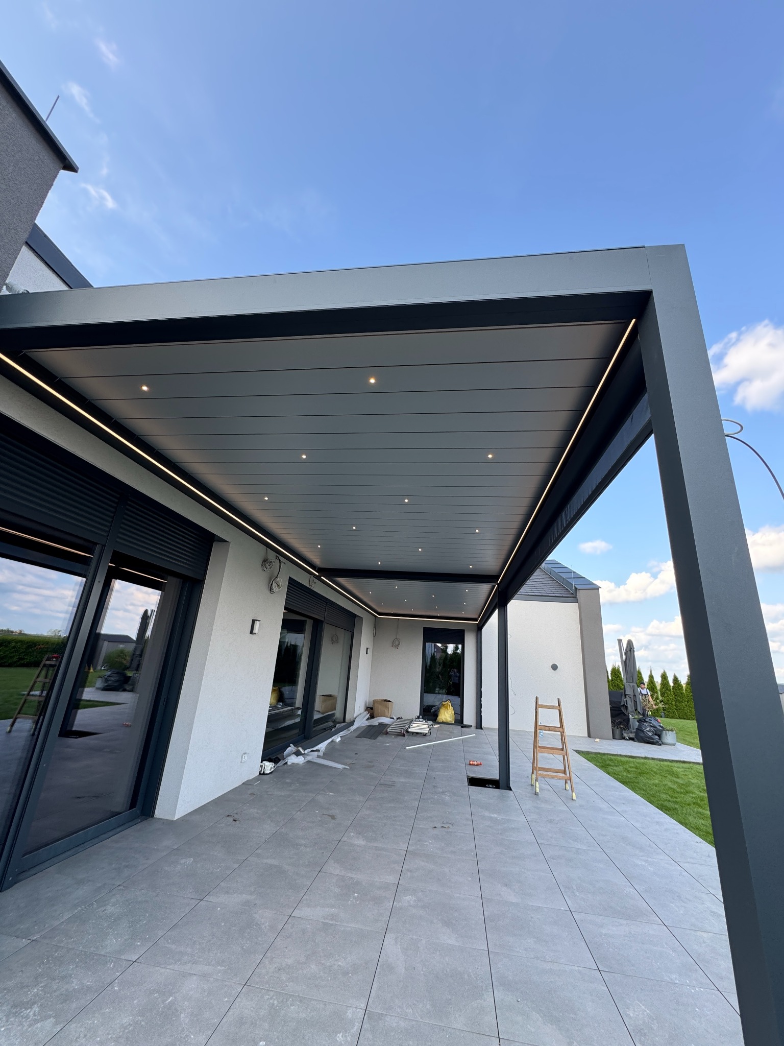 Nowoczesna pergola tarasowa z oświetleniem LED, widok z dołu, szare płytki na podłodze, częściowo widoczne okna i zieleń w tle, błękitne niebo.
