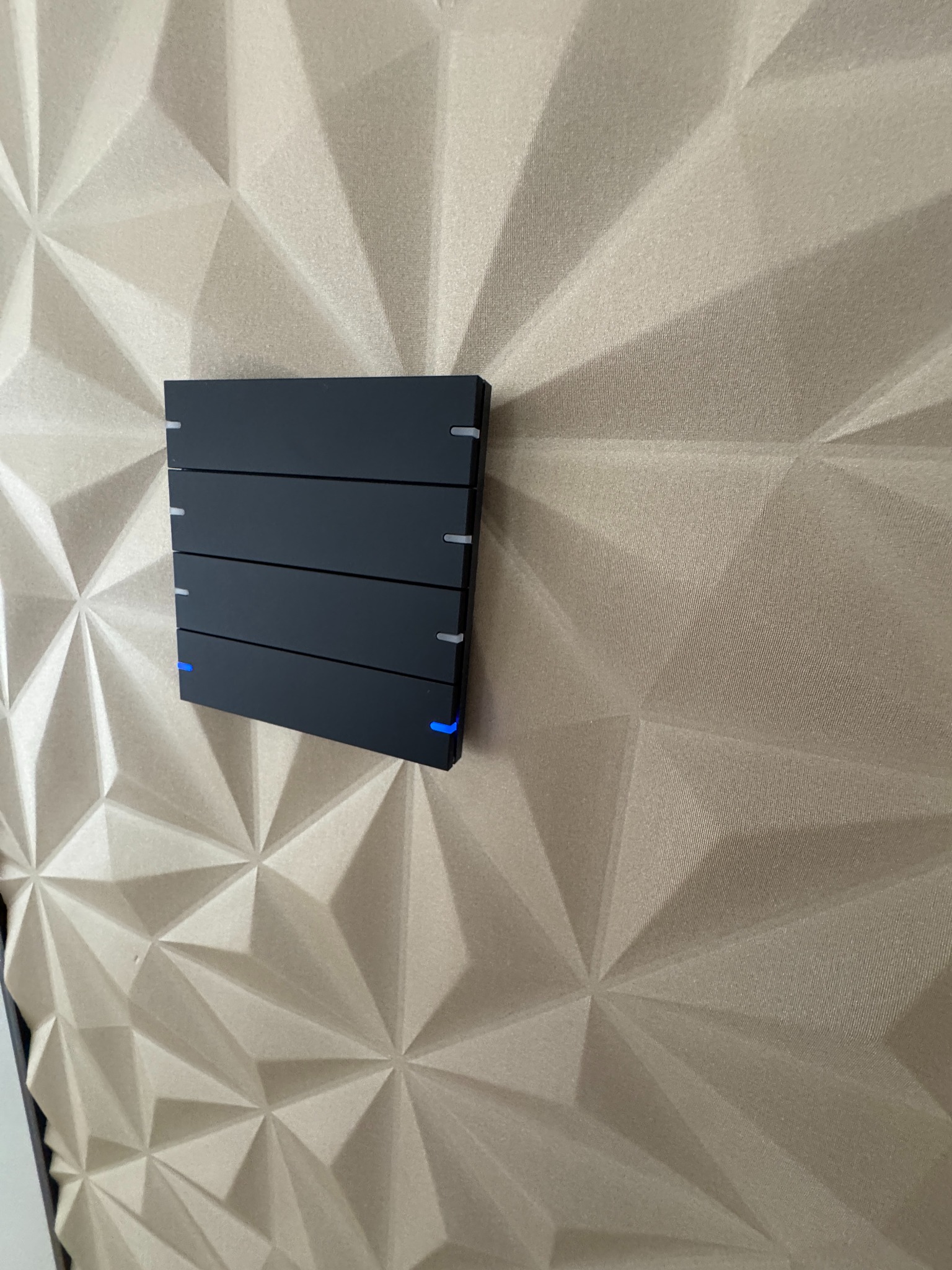 Czarny panel sterowania inteligentnego domu z subtelnym, niebieskim podświetleniem LED, zamontowany na dekoracyjnej ścianie z trójwymiarową fakturą.