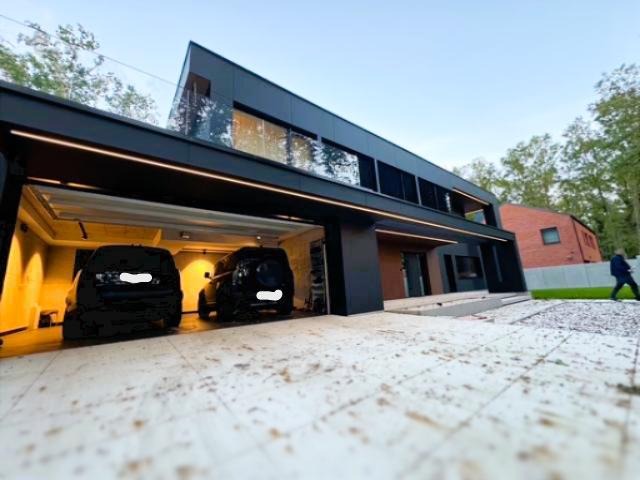 Nowoczesny dom z garażem, oświetleniem LED i dwoma samochodami. Widok z dołu, podkreślający architekturę i oświetlenie zewnętrzne.