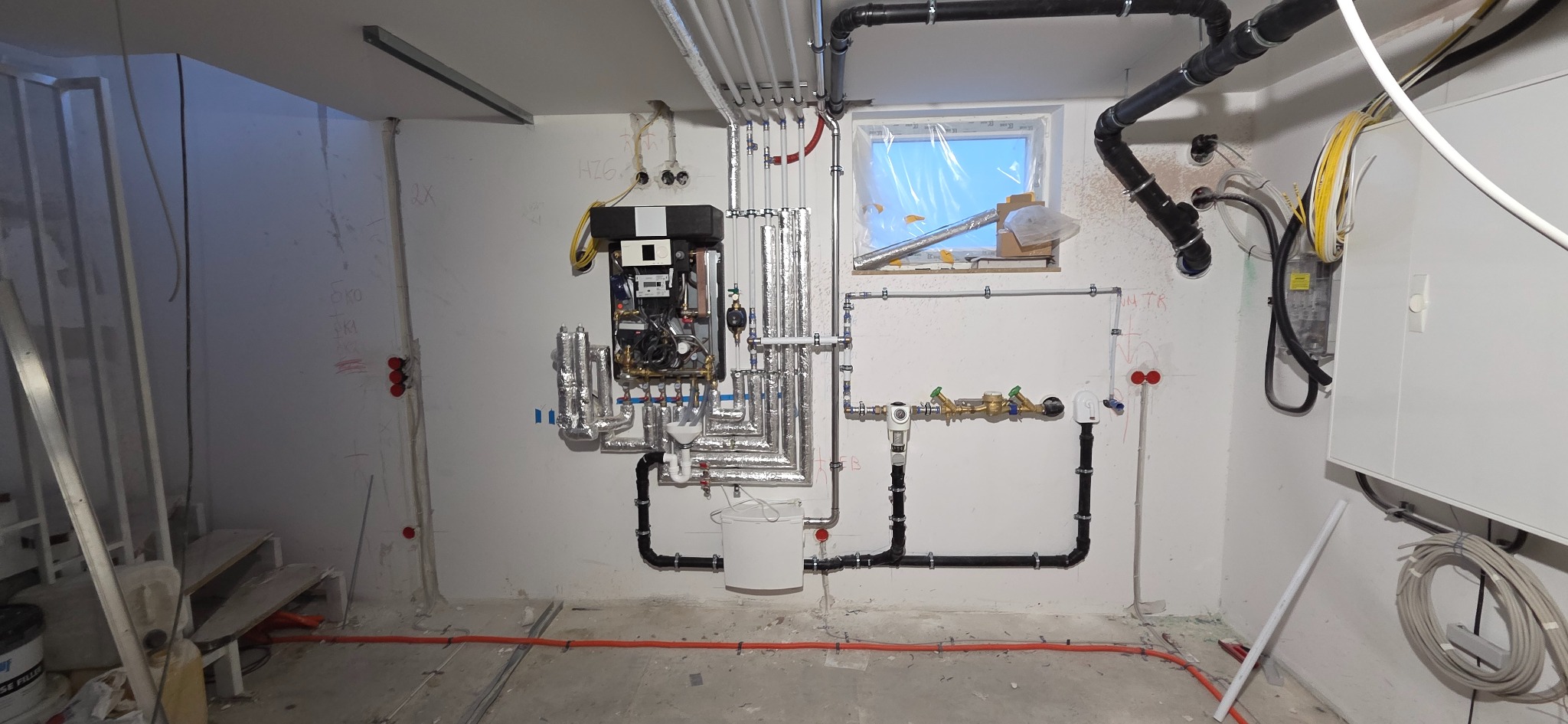 Złożona instalacja grzewcza z kotłem, rurami w izolacji i elementami armatury, zamontowana na ścianie w surowym pomieszczeniu z oknem i widocznymi instalacjami elektrycznymi.