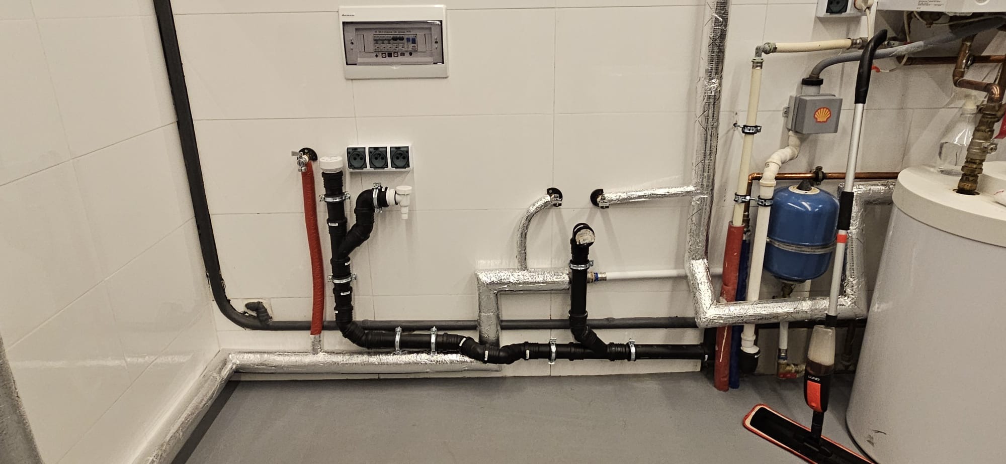 Złożona instalacja hydrauliczna z rurami w izolacji, zaworami i zbiornikiem, zamontowana na białej, kafelkowanej ścianie w pomieszczeniu technicznym. Widoczna skrzynka elektryczna.