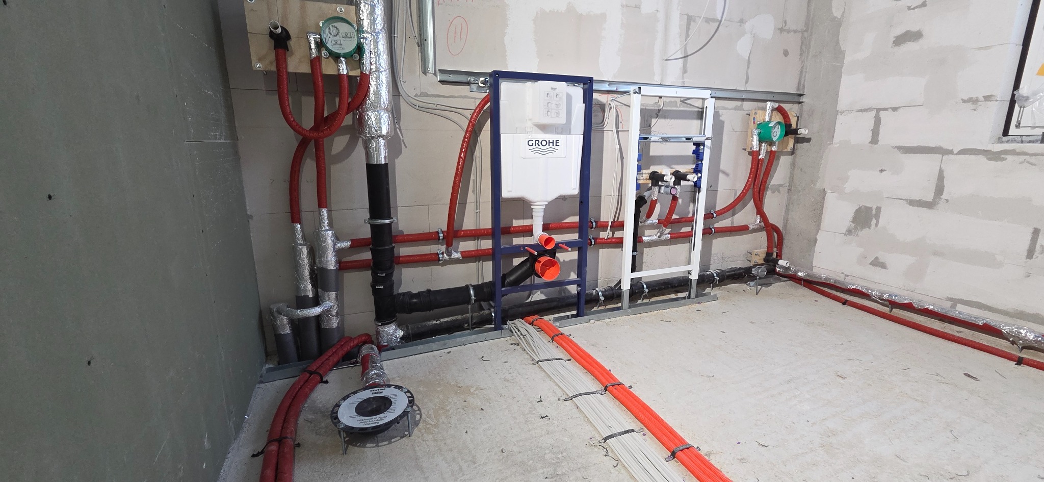 Instalacja wodno-kanalizacyjna w surowym stanie: rury, stelaż podtynkowy Grohe, liczniki. Czerwone rury grzewcze, czarne rury kanalizacyjne, szare ściany w tle.