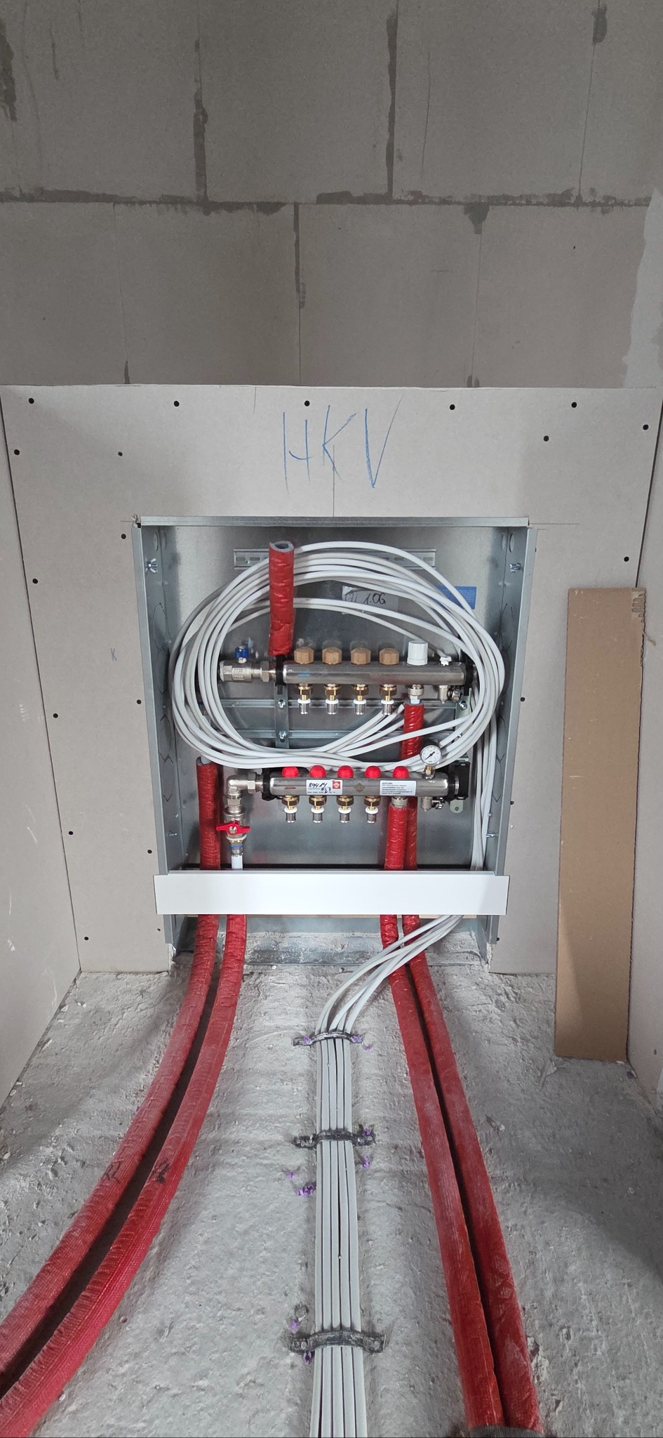Rozdzielacz ogrzewania podłogowego w metalowej szafce, z czerwonymi rurami w podłodze i białymi kablami. Widoczne napisy HKV na ścianie z płyt kartonowo-gipsowych.