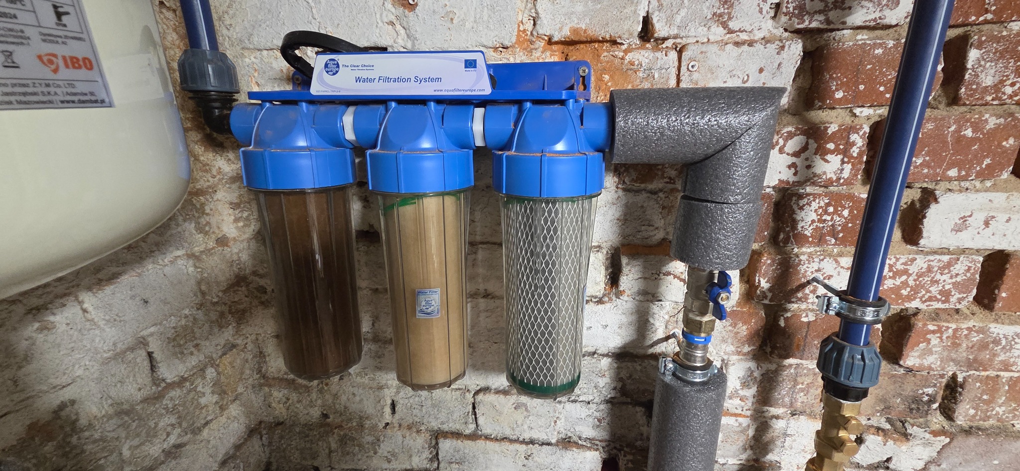 System filtracji wody z trzema przezroczystymi filtrami o różnym stopniu zabrudzenia, zamontowany na ceglanej ścianie. Widoczne rury i zawory.