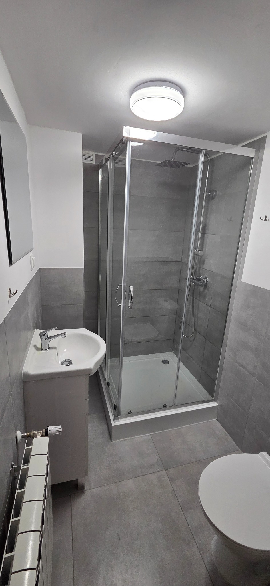 Nowoczesna łazienka z kabiną prysznicową typu walk-in, umywalką z szafką i grzejnikiem. Szare płytki na ścianach i podłodze. Minimalistyczny design.
