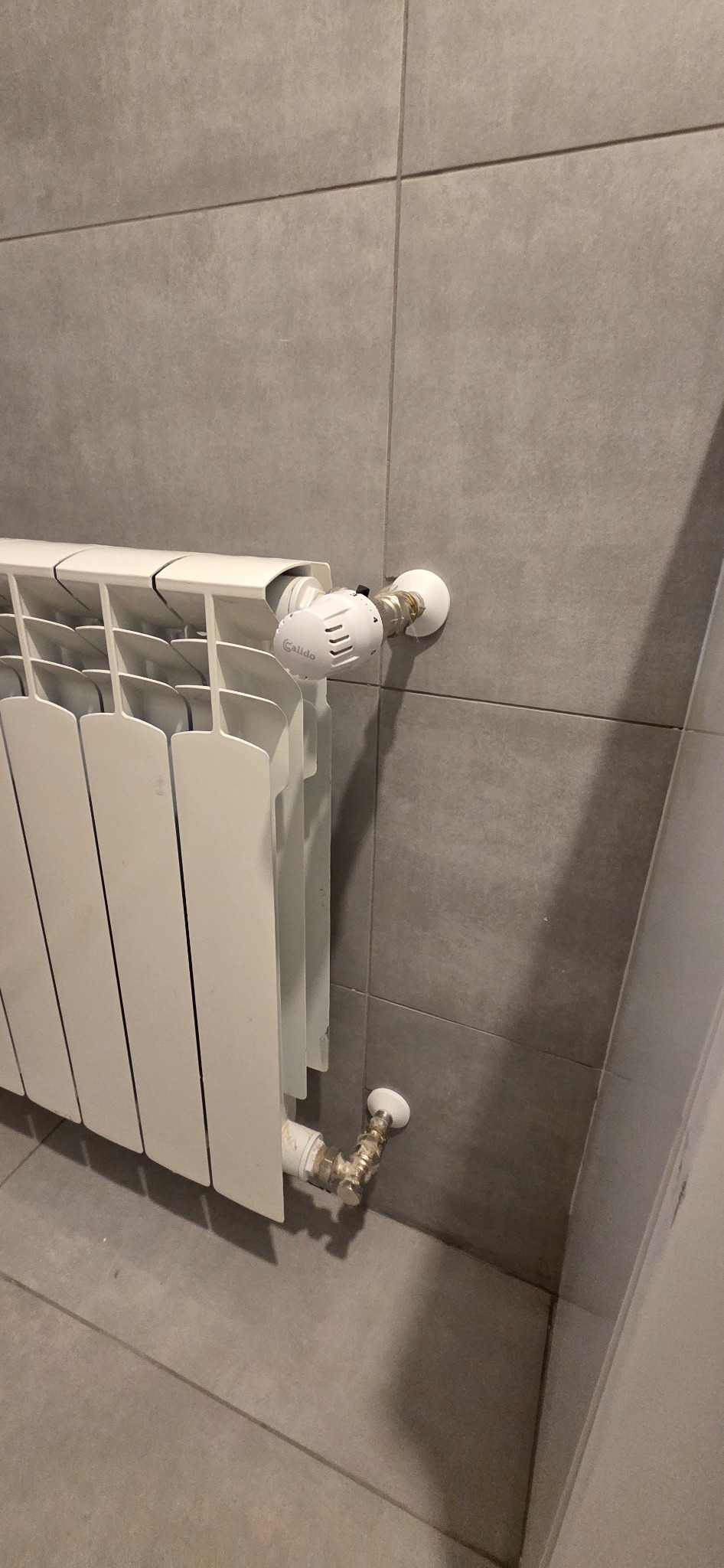 Biały grzejnik aluminiowy z termostatem Calido na szarej, kwadratowej płytce ściennej i podłogowej. Widoczne przyłącza hydrauliczne.