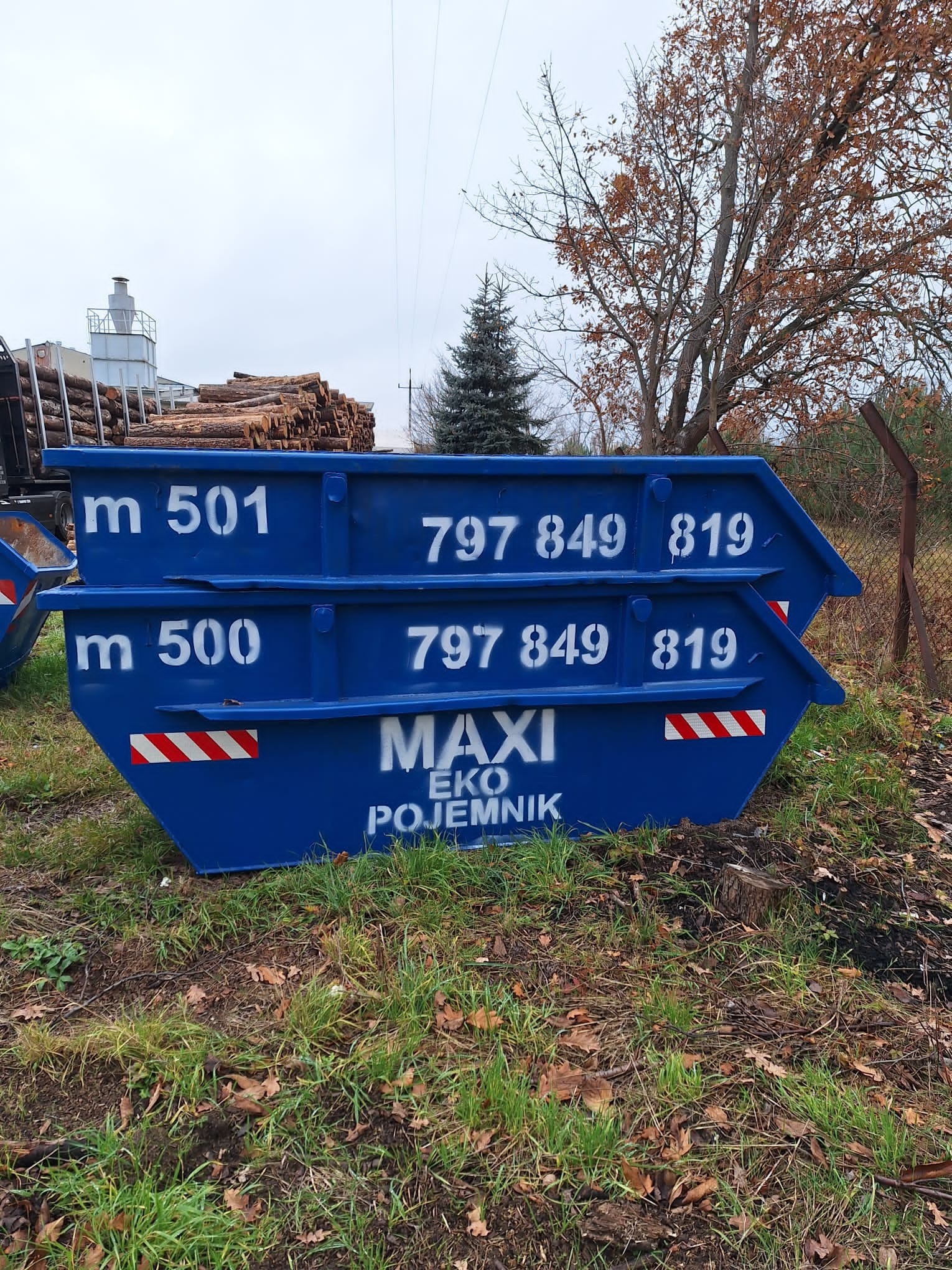 Dwa niebieskie, puste kontenery MAXI EKO POJEMNIK, ustawione jeden na drugim, z numerami 500 i 501, na tle trawy i drzew. Widoczny załadunek drewna w tle.