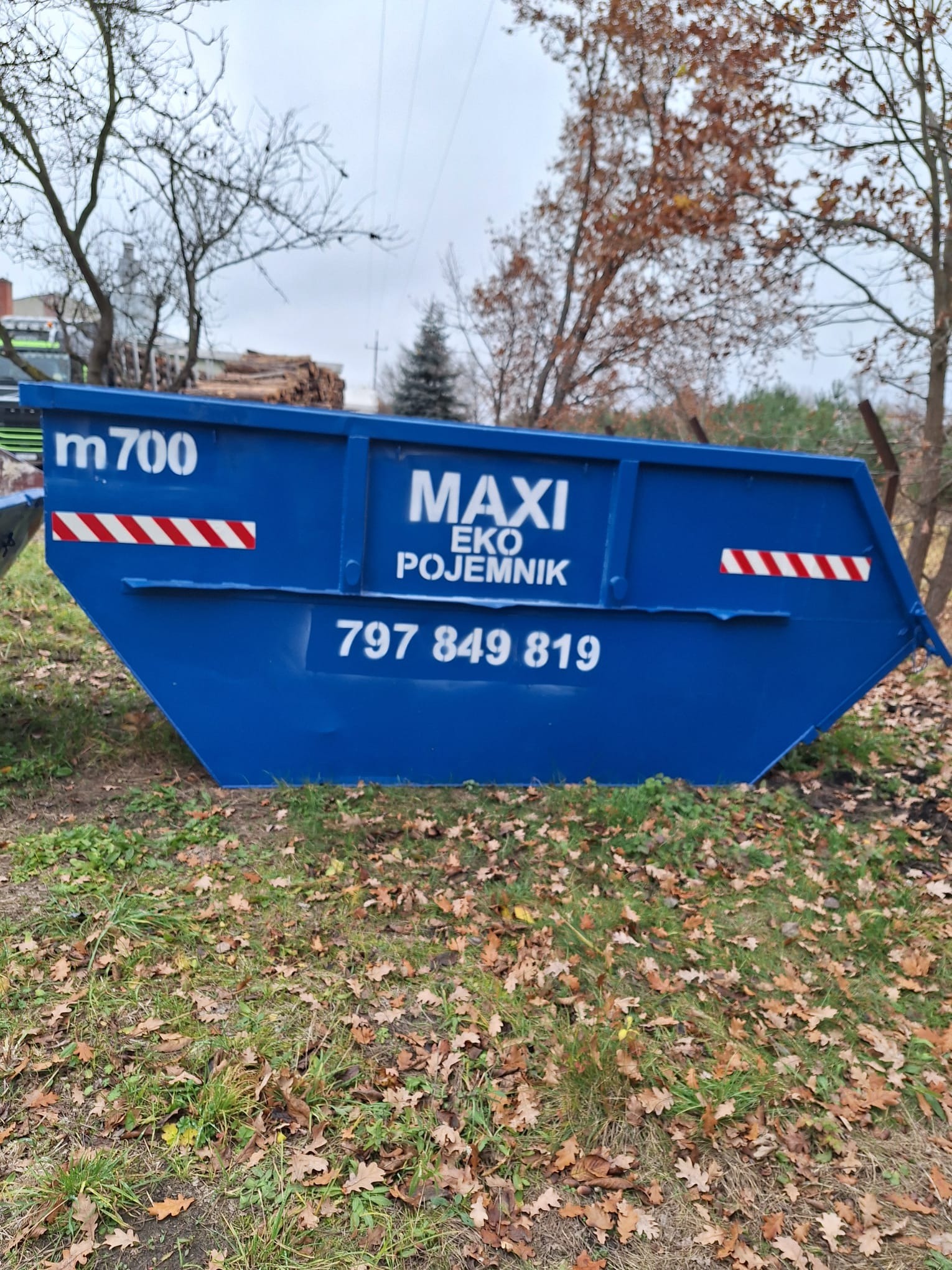 Niebieski kontener z napisem 'MAXI EKO POJEMNIK' i numerem telefonu, stojący na trawie z liśćmi. Widoczne drzewa w tle, pochmurne niebo.
