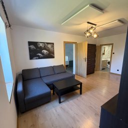 Golden Doors Nieruchomości Alan Fiszebrandt - Jasne wnętrze mieszkania z szarą sofą narożną, czarnym stolikiem kawowym i grafiką z meduzami na ścianie. Widoczne drzwi do innych pomieszczeń i drewniana podłoga.