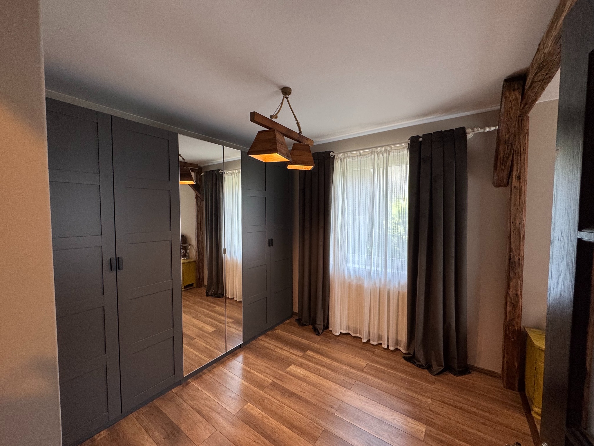 Nowoczesna sypialnia w Bielsku-Białej z drewnianą podłogą, szarą szafą wnękową z lustrem i designerską lampą. Widoczne drewniane elementy konstrukcyjne.