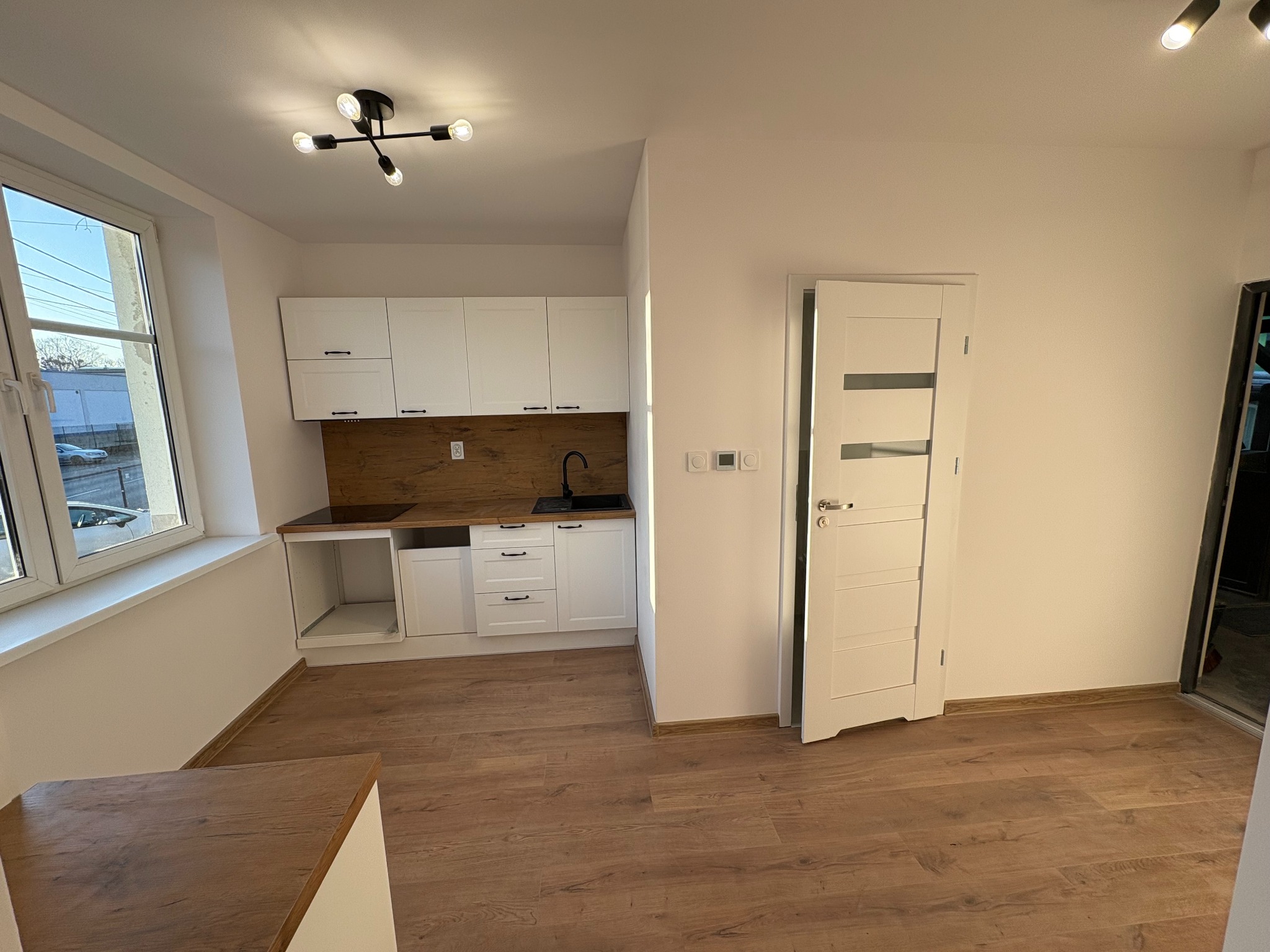 Jasne wnętrze: biała kuchnia z drewnianym blatem i podłogą, nowoczesna lampa sufitowa, okno z widokiem na zewnątrz, białe drzwi z przeszkleniami.