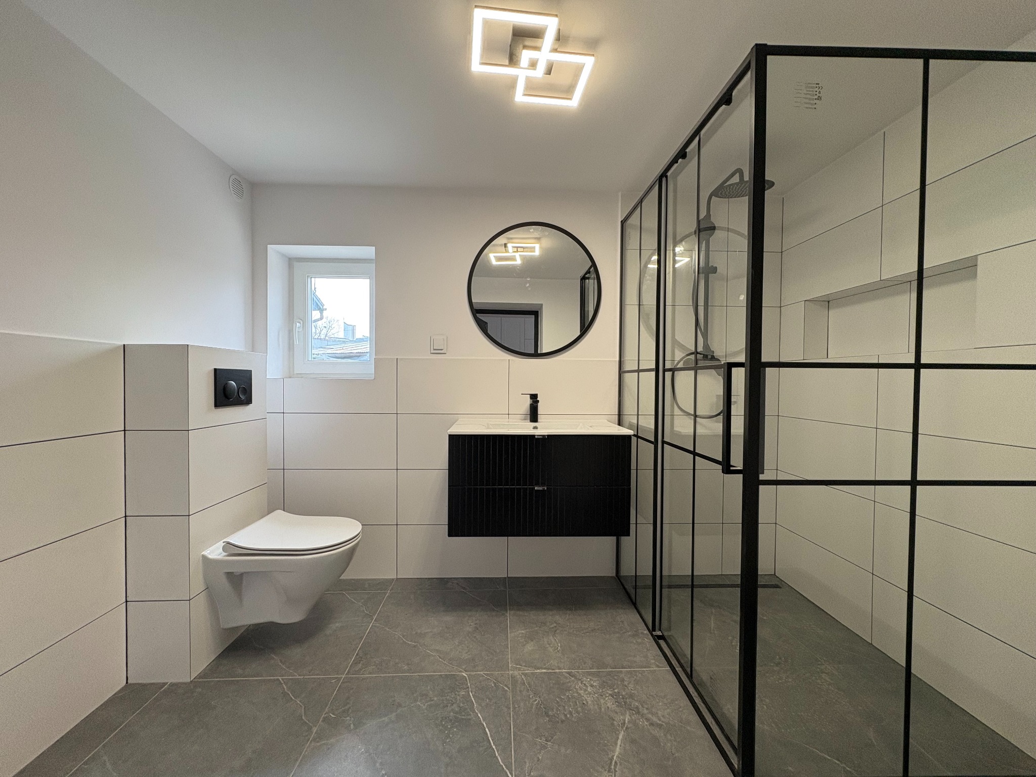 Nowoczesna łazienka z kabiną prysznicową typu walk-in, wiszącą toaletą i czarną szafką pod umywalką. Szare płytki podłogowe imitujące marmur, białe płytki ścienne.