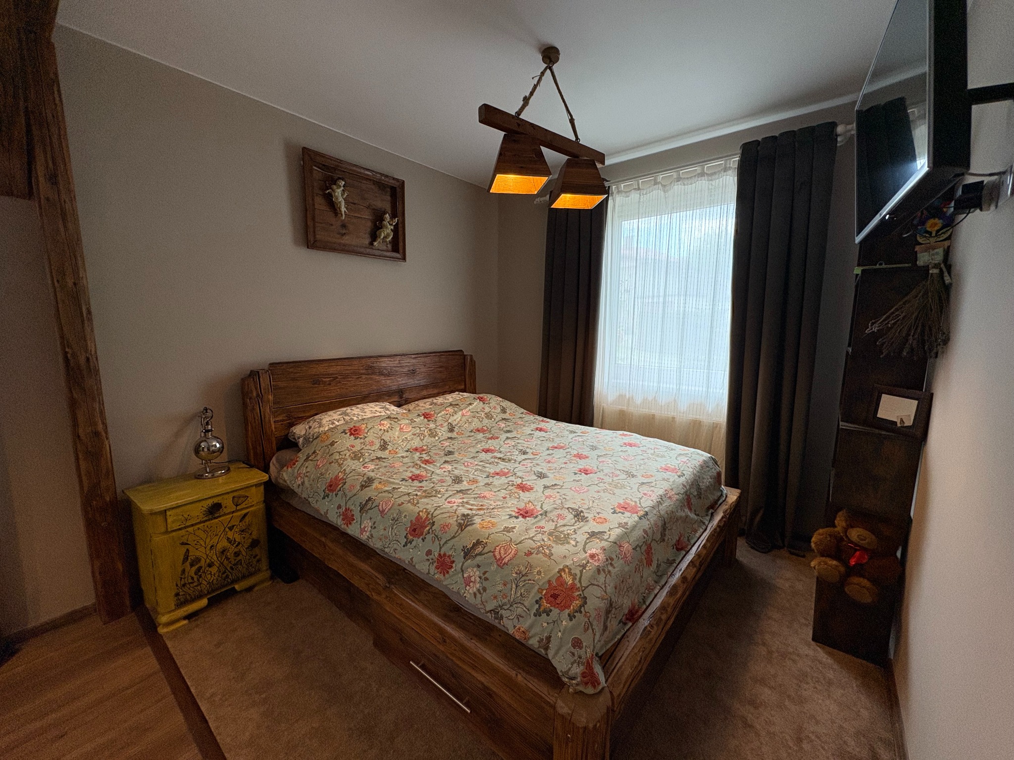 Rustykalna sypialnia z drewnianym łóżkiem i szafką nocną w odcieniach żółci. Na ścianie dekoracje i telewizor. Przytulne wnętrze z charakterem.