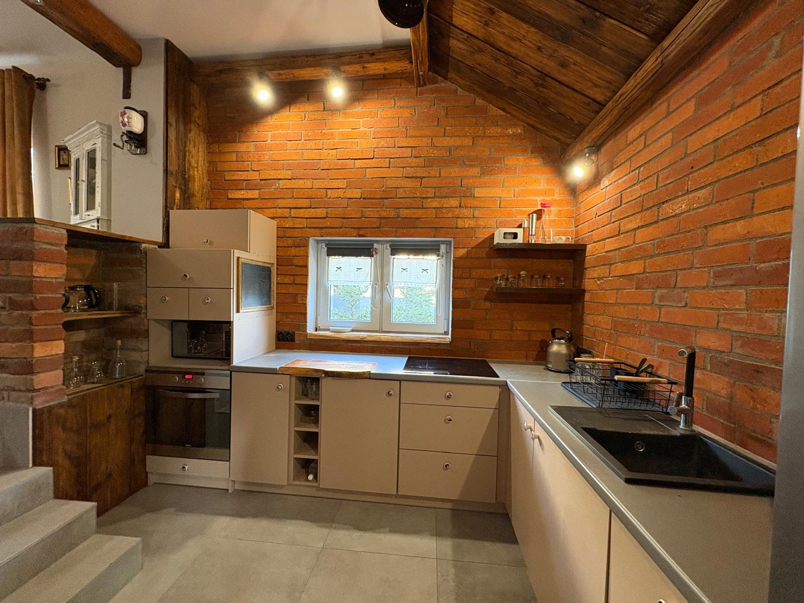 Rustykalna kuchnia z ceglaną ścianą, drewnianym stropem i beżowymi szafkami. Widoczne schody, okno z widokiem na zieleń i czarny zlew. Oświetlenie punktowe.