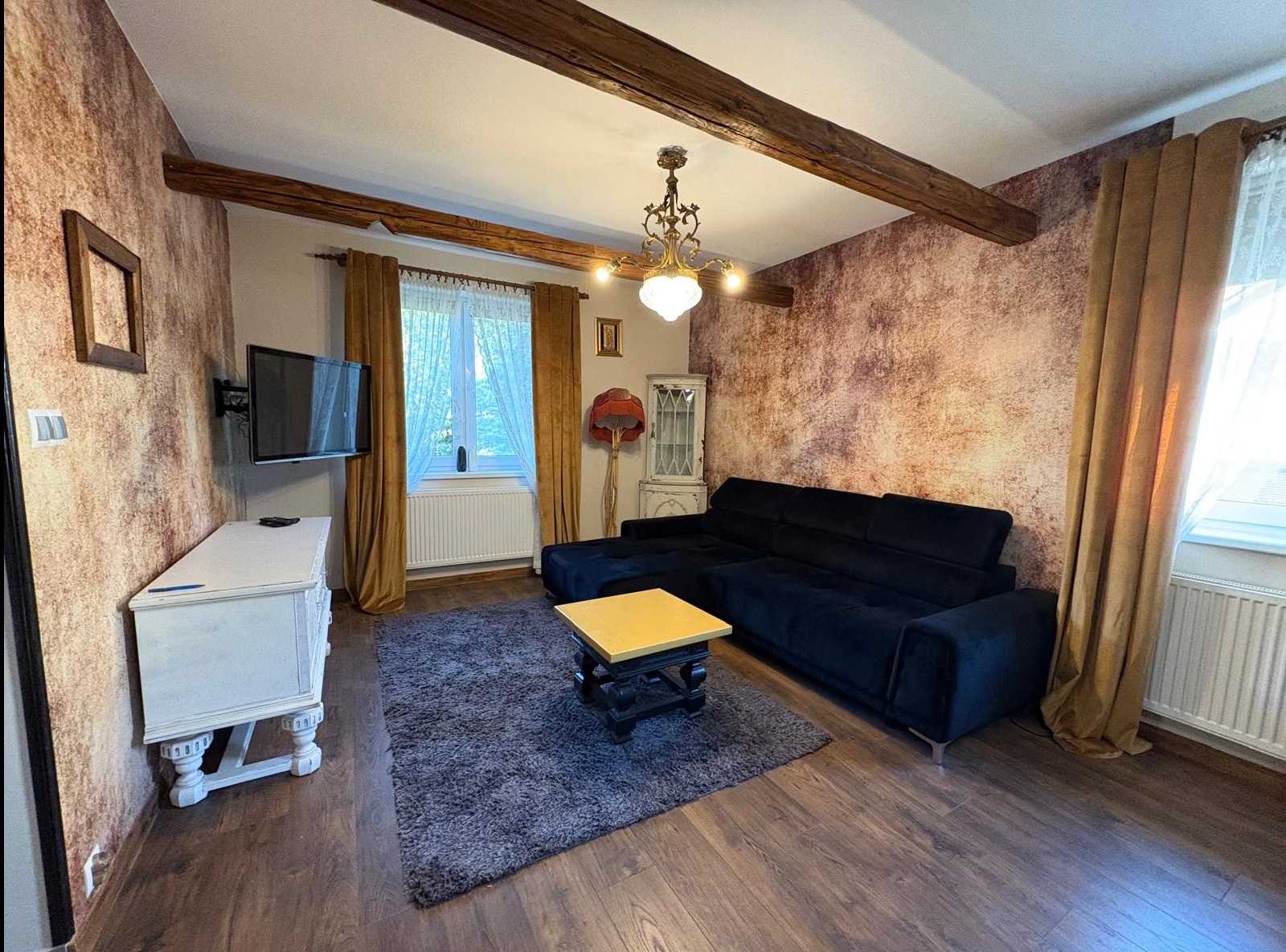 Salon z czarną sofą narożną, drewnianymi belkami stropowymi i wzorzystą tapetą. Centralnie dywan i stolik kawowy. Widok na okno z zasłonami.