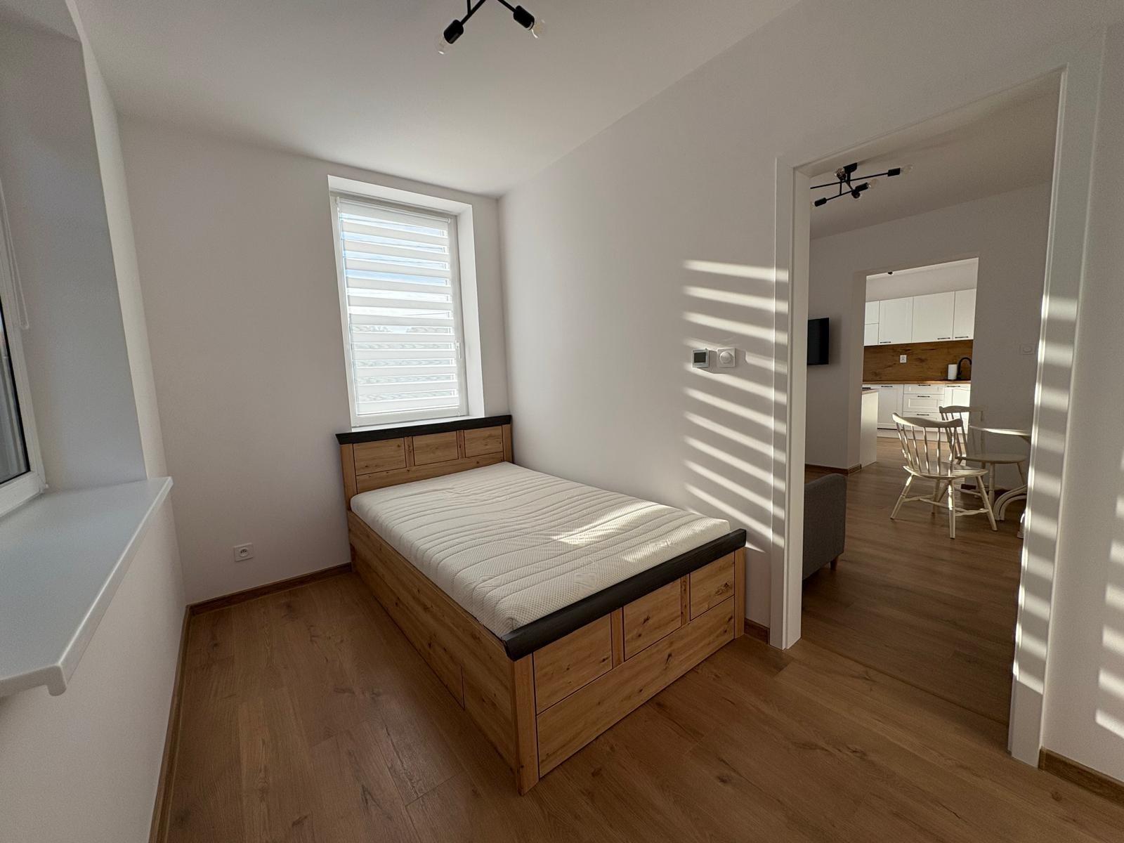 Jasny pokój z drewnianym łóżkiem i widokiem na kuchnię przez otwarte drzwi. Promienie słońca wpadają przez żaluzje, oświetlając podłogę i ścianę.