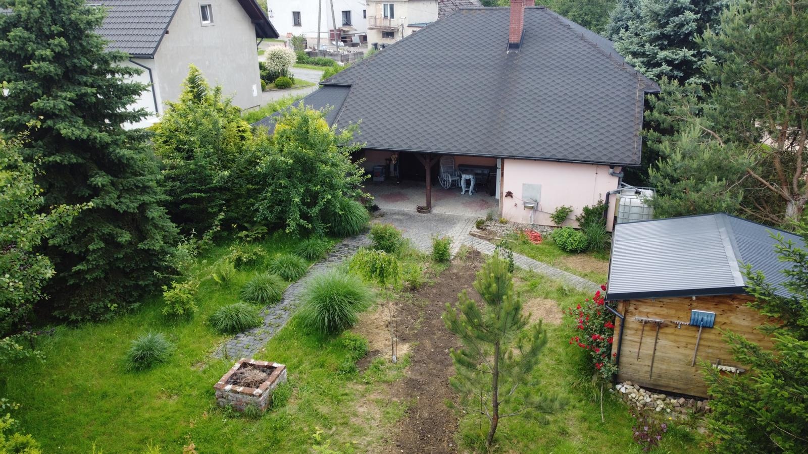 Dom z ogrodem z perspektywy lotu ptaka. Widoczny budynek z ciemnym dachem, zielony ogród z kamienną ścieżką i drewniana szopa na narzędzia. Ujęcie z góry.