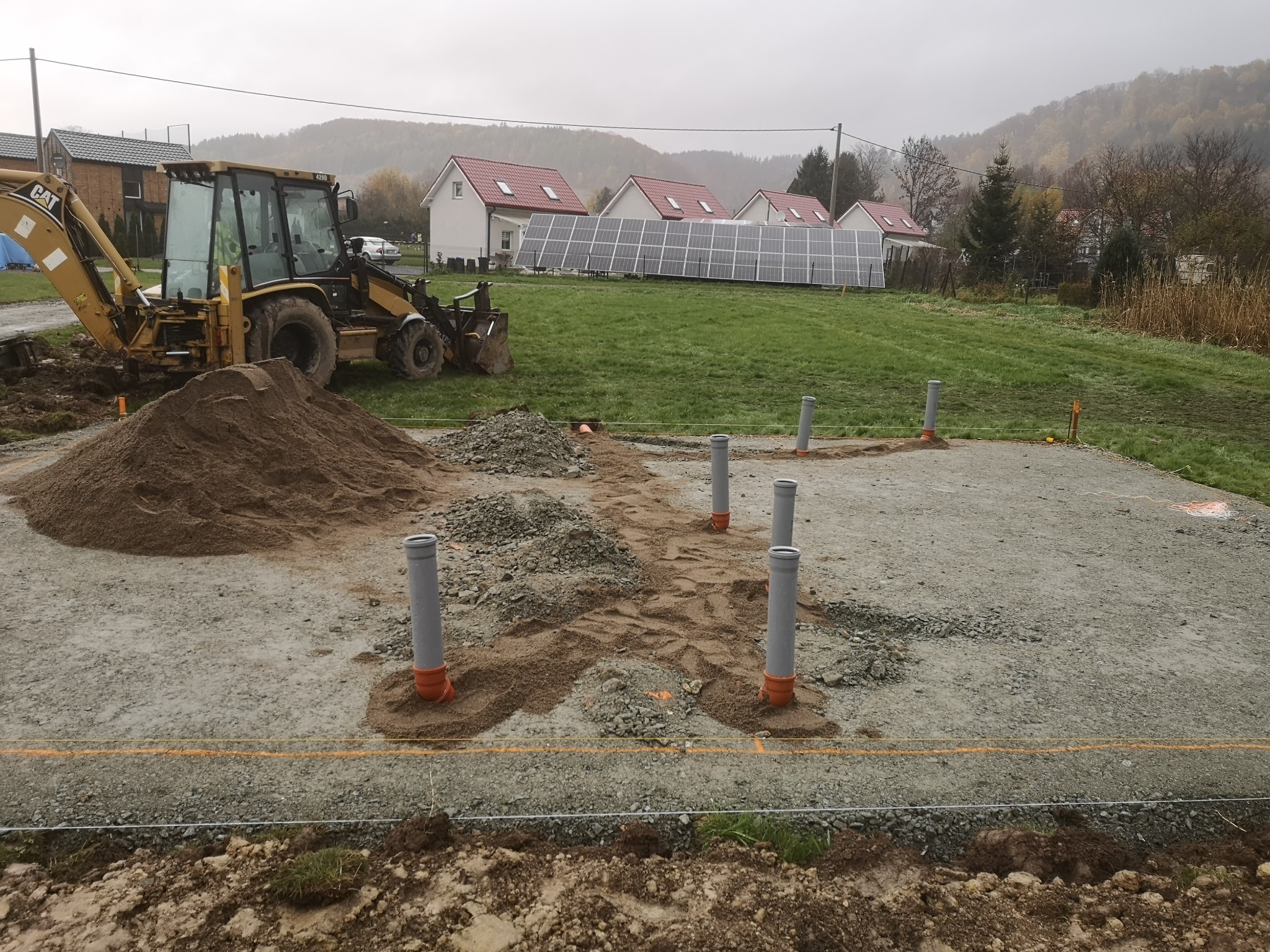 Żółta koparko-ładowarka CAT obok pryzmy piasku na przygotowanym terenie pod budowę. Widoczne pionowe rury kanalizacyjne i panele słoneczne w tle.