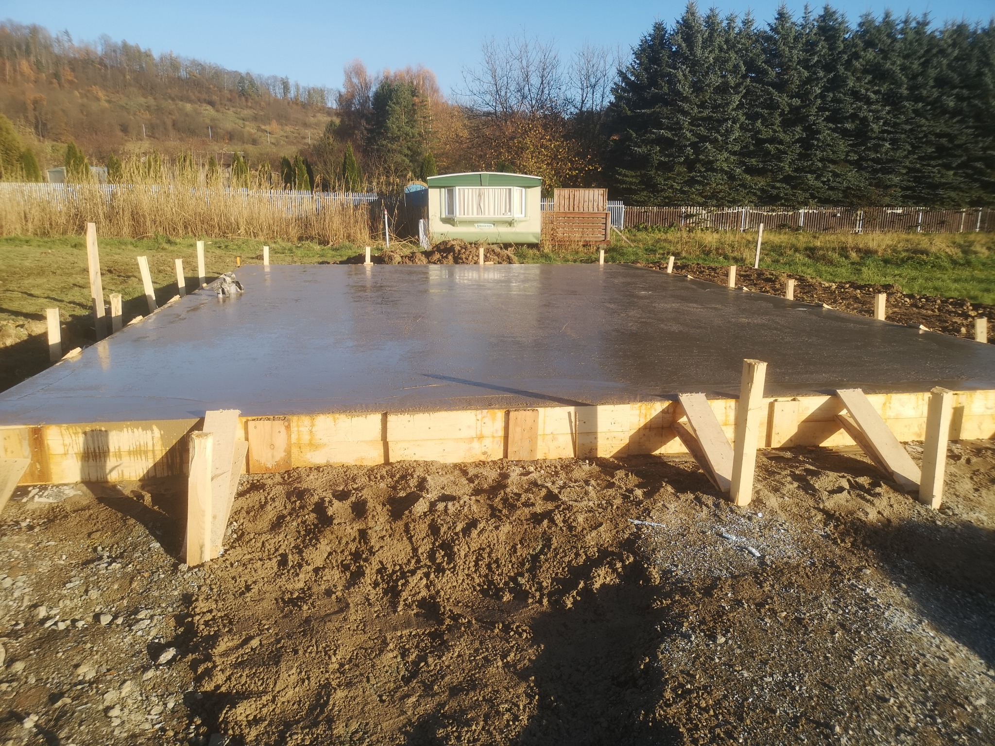 Zalany fundament pod budynek z drewnianym szalunkiem w słoneczny dzień. W tle przyczepa kempingowa i drzewa. Widok na świeży beton i przygotowany teren budowy.