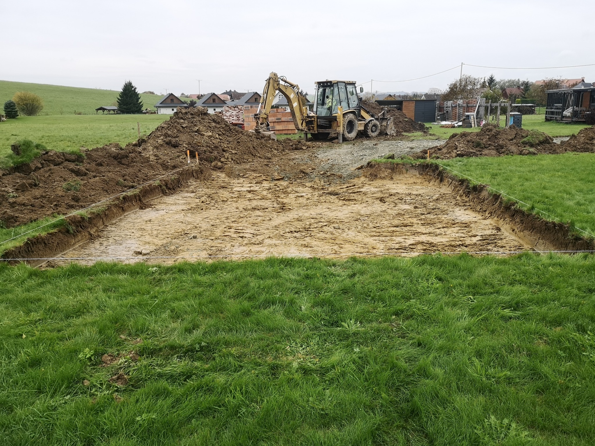 Wykop pod fundamenty z żółtą koparką w tle, widok z poziomu gruntu. Wykopane obrzeża wyznaczone sznurkiem. W tle zabudowa jednorodzinna i zieleń.