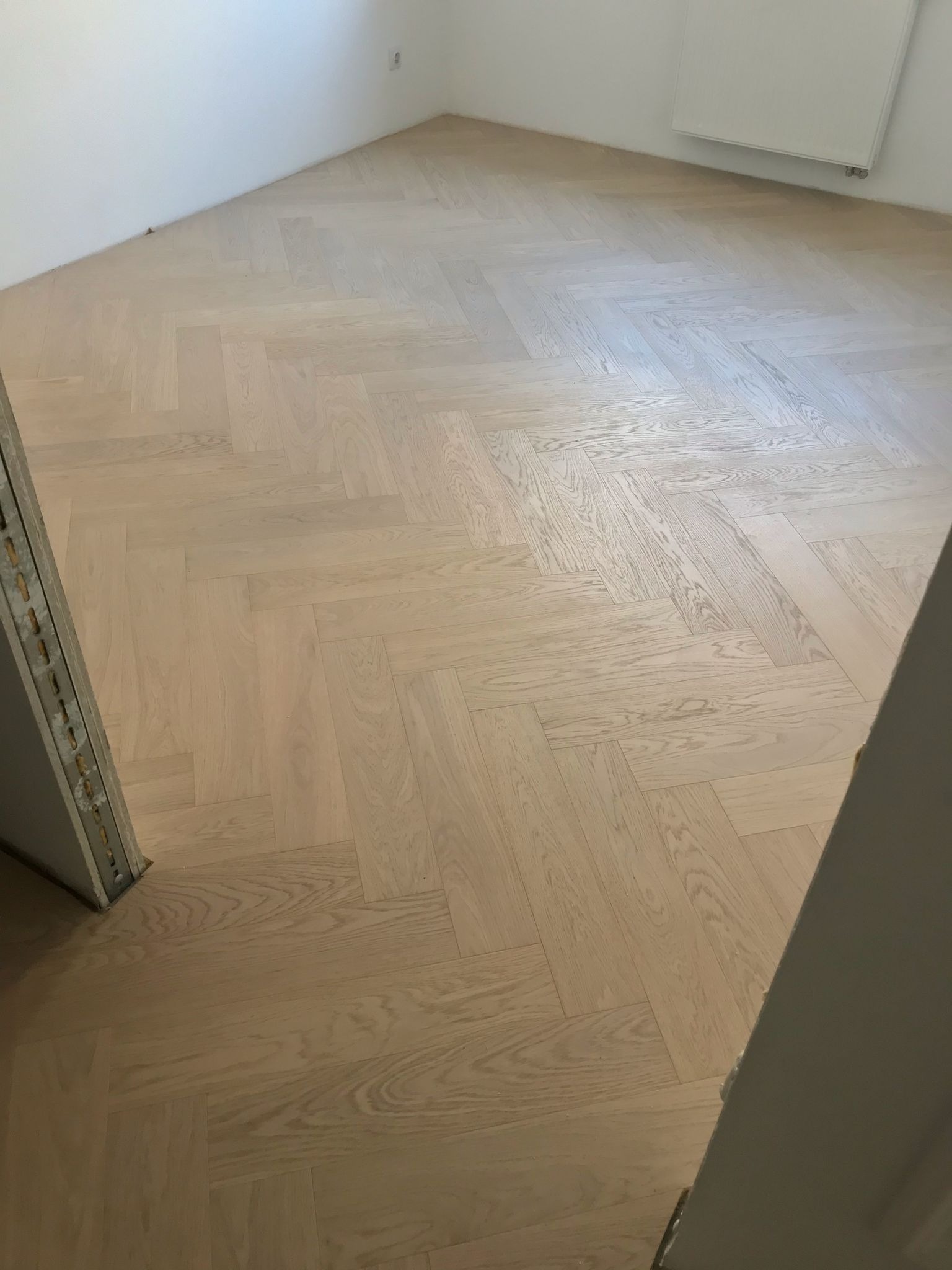 Jasna, nowa podłoga ułożona w jodełkę francuską, widoczna z perspektywy wejścia do pokoju. Świeżo wykończone wnętrze z białymi ścianami i grzejnikiem.