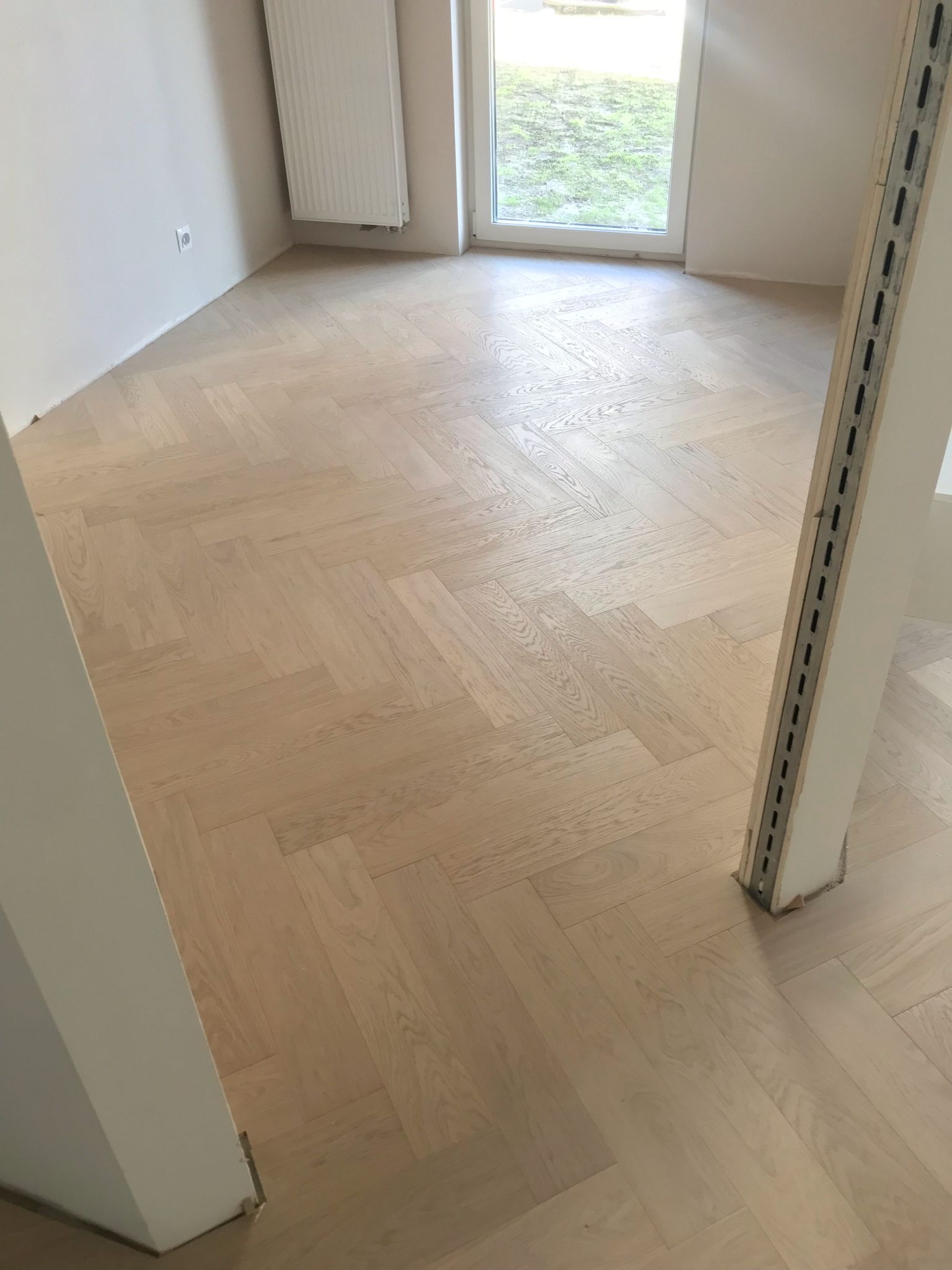 Jasny parkiet ułożony we wzór jodełki francuskiej w nowym wnętrzu. Widoczne detale wykończenia przy ścianach i oknie. Minimalistyczny styl.