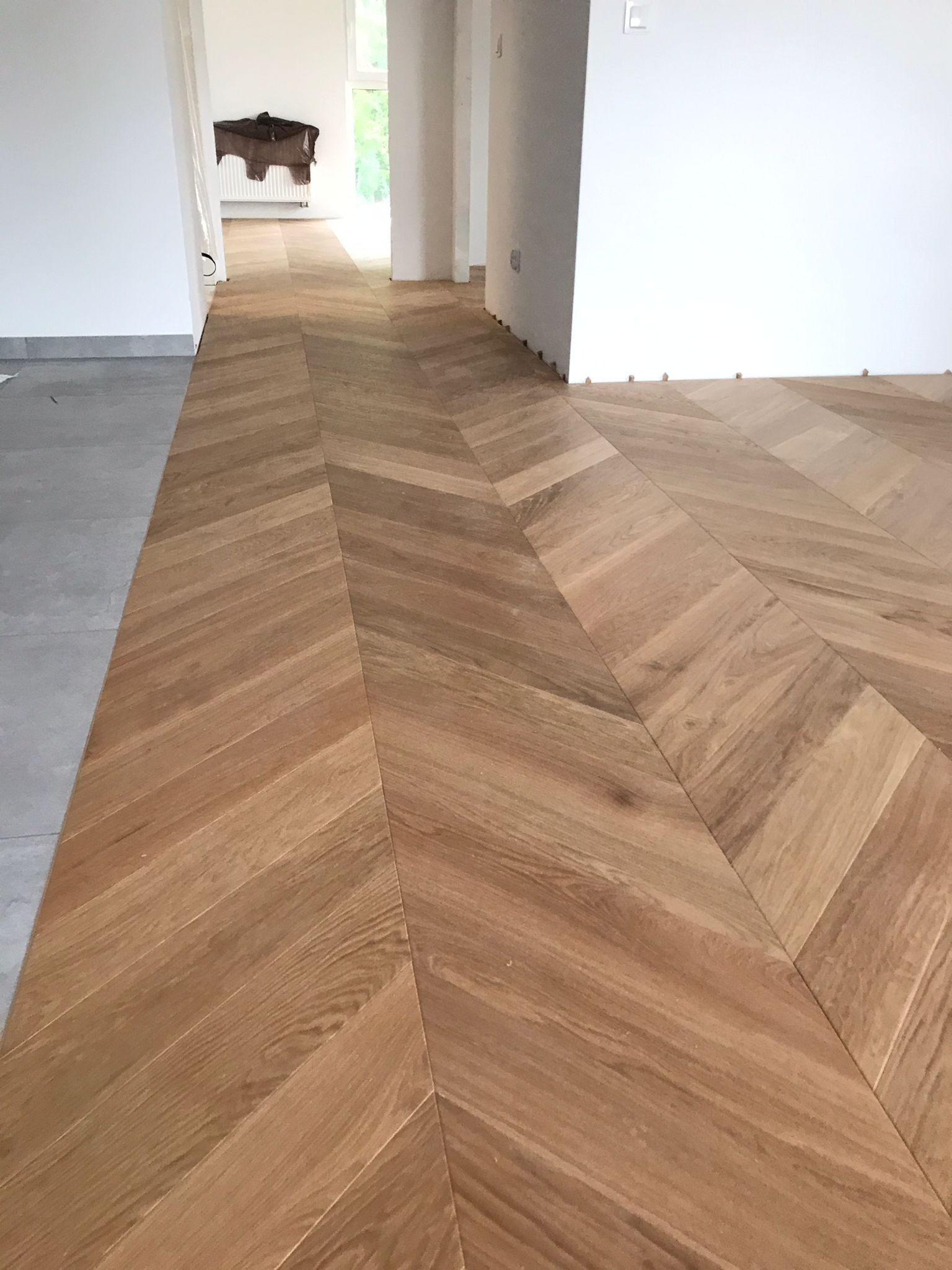 Ułożony parkiet w jodełkę w jasnym wnętrzu. Widoczne listwy przypodłogowe i fragment szarej podłogi w przejściu. Naturalne oświetlenie.