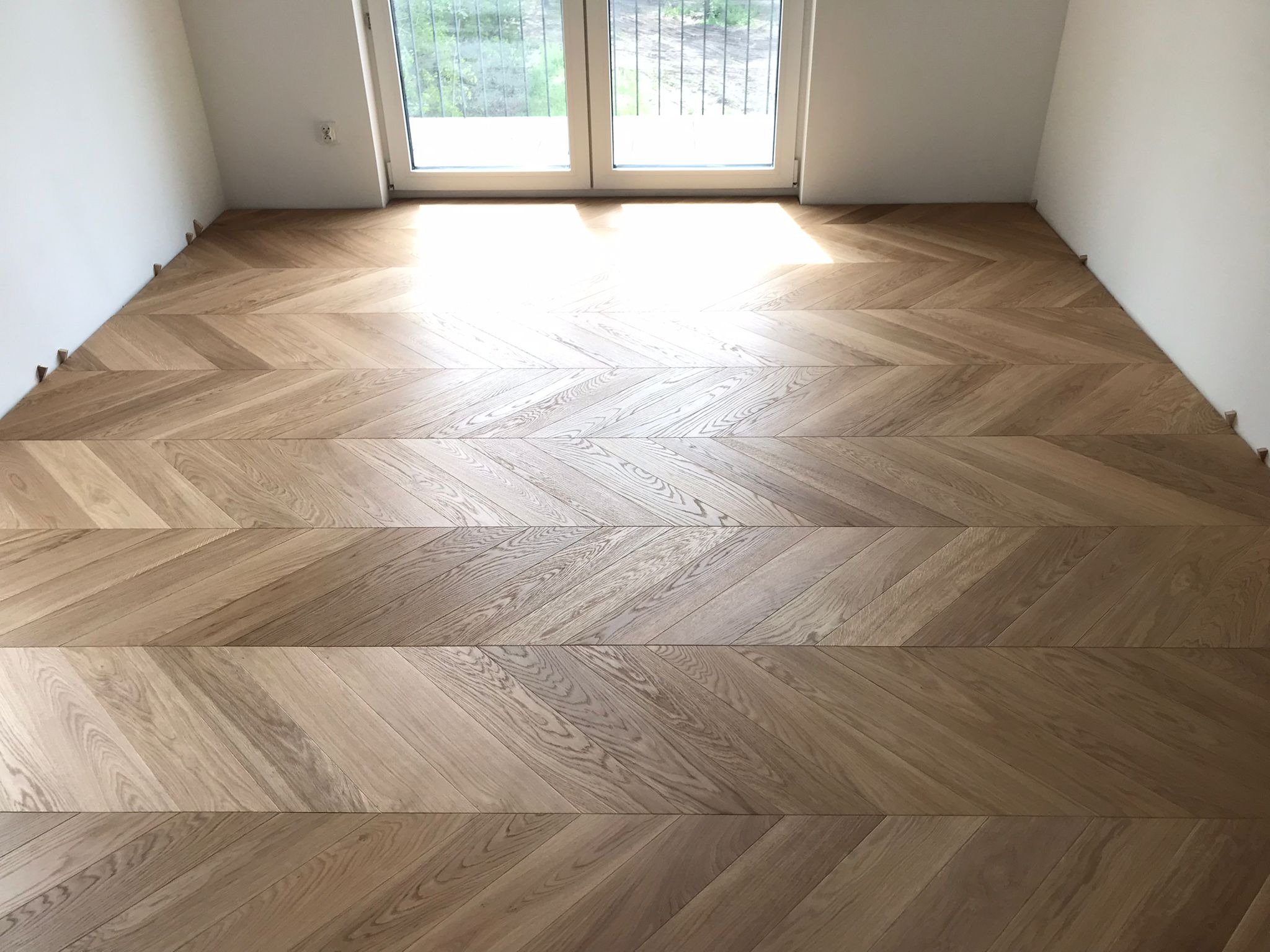 Jasna, drewniana podłoga ułożona we wzór jodełki francuskiej, z widocznymi klinami dystansowymi przy ścianach i oknem w tle, w minimalistycznym wnętrzu.