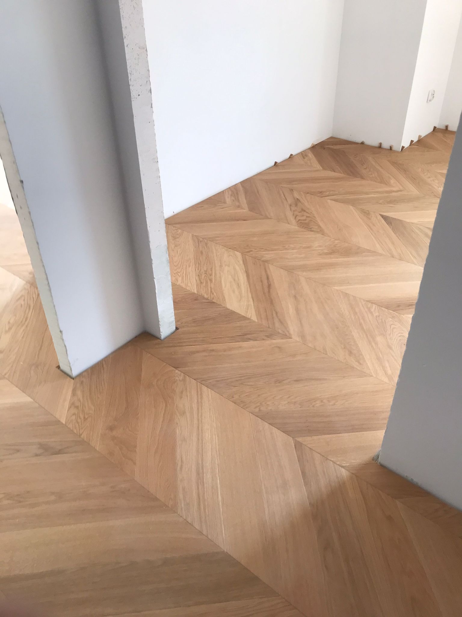 Ułożony parkiet w jodełkę francuską, jasne drewno, widoczne szczeliny dylatacyjne przy ścianach, surowy stan wykończenia wnętrza, białe ściany.