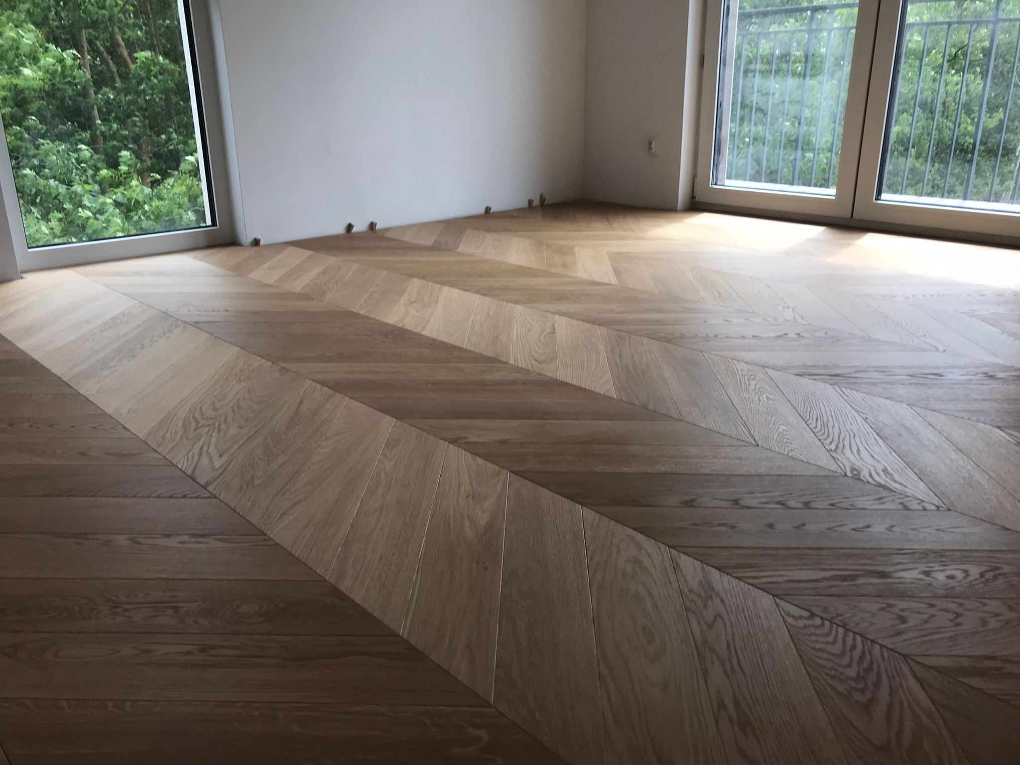 Jasny parkiet ułożony we wzór jodełki francuskiej w przestronnym pokoju z dużymi oknami. Widoczne drewniane kołki dystansowe przy ścianie.