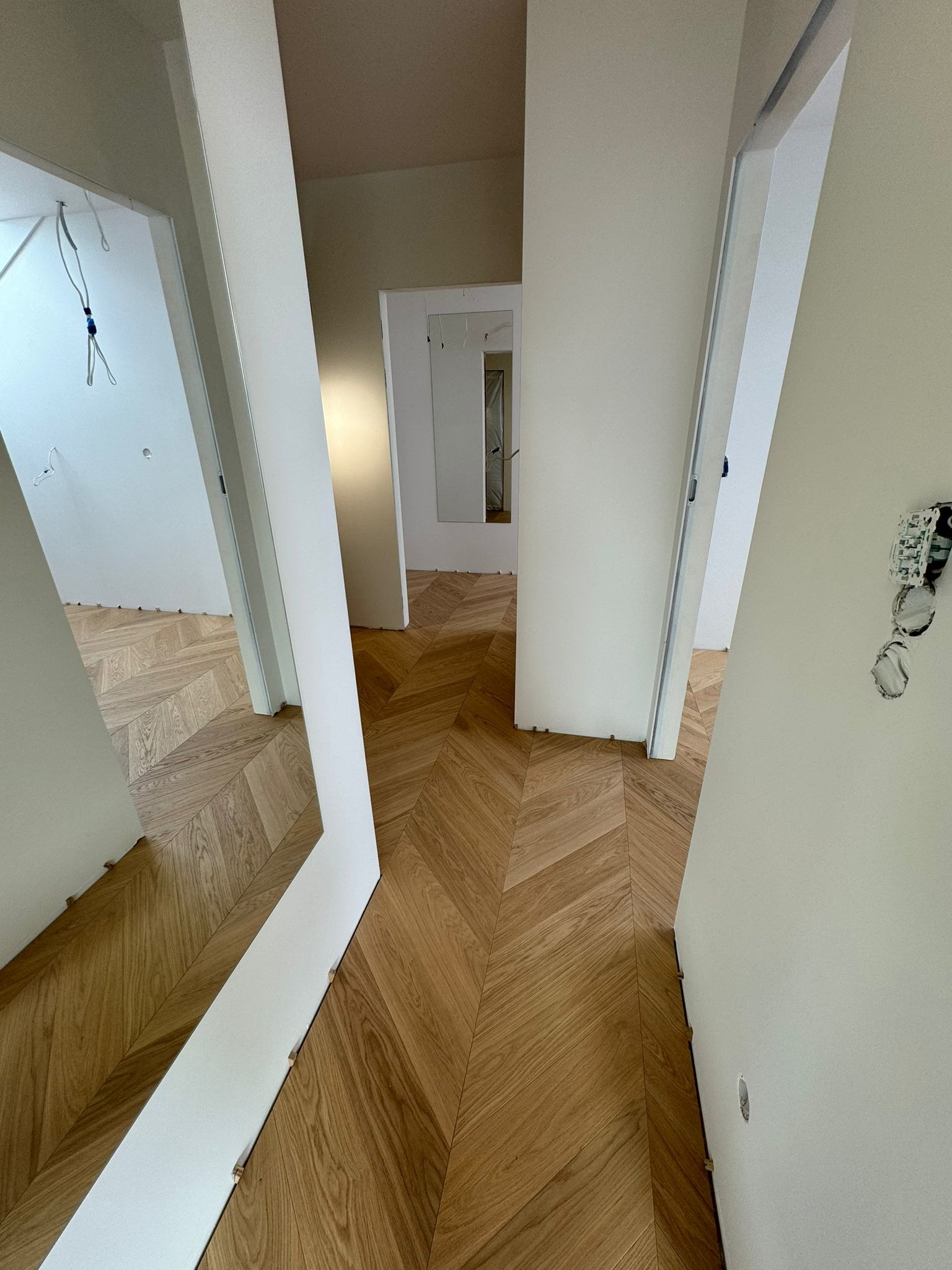 Ułożony parkiet w jodełkę francuską w jasnym korytarzu z białymi ścianami i otwartymi drzwiami. Widoczne niedokończone gniazdko elektryczne.