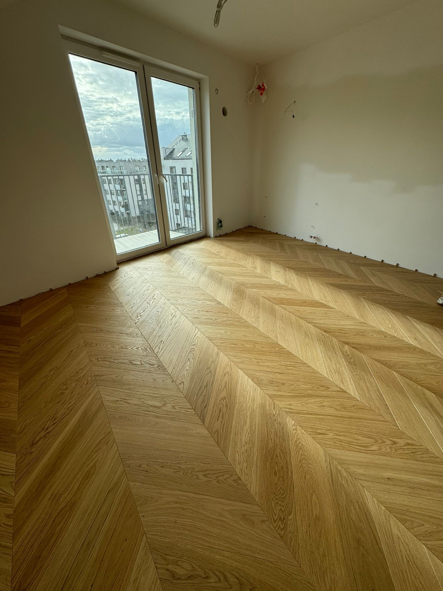 Ukończona podłoga z parkietu ułożonego w jodełkę w jasnym pomieszczeniu z dużym oknem balkonowym i widokiem na miasto. Widoczne listwy dystansowe przy ścianach.