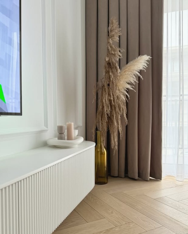 Jasna jodełka klasyczna na podłodze, uzupełniona o białą szafkę RTV z ryflowanym frontem i dekoracjami w postaci traw pampasowych i świec.