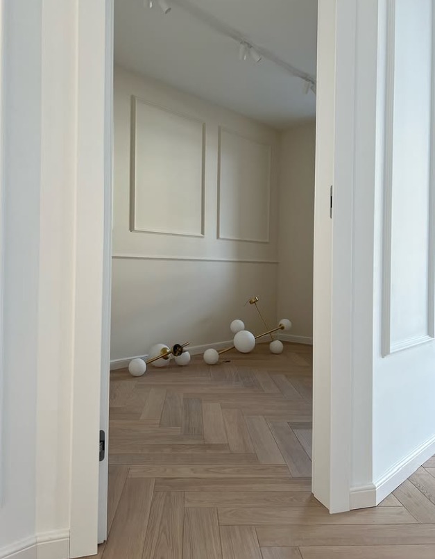 Jasny parkiet ułożony we wzór jodełki francuskiej, widoczny zza otwartych, białych drzwi. W tle minimalistyczna lampa i ściany z dekoracyjnymi panelami.