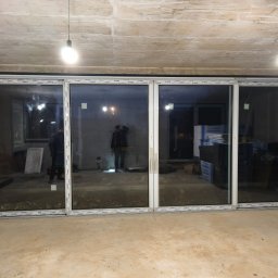 Perfect Montaż Roman Tselikov - Czteroskrzydłowe drzwi przesuwne z białymi profilami Aluplast, zamontowane w surowym wnętrzu z betonowym sufitem i podłogą. Widoczne odbicia osób w szybach.