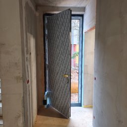 Perfect Montaż Roman Tselikov - Otwarta, szara, zewnętrzna aluminiowa drzwi z szybą, z widocznym logo producenta, w surowym wnętrzu budynku w trakcie wykończenia. Widok na zewnątrz.