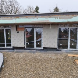 Perfect Montaż Roman Tselikov - Montaż okien PCV na etapie wykończenia elewacji budynku. Widoczne ocieplenie, surowa ściana z naklejonym styropianem i trzy okna z białymi ramami. Na dole żwir.