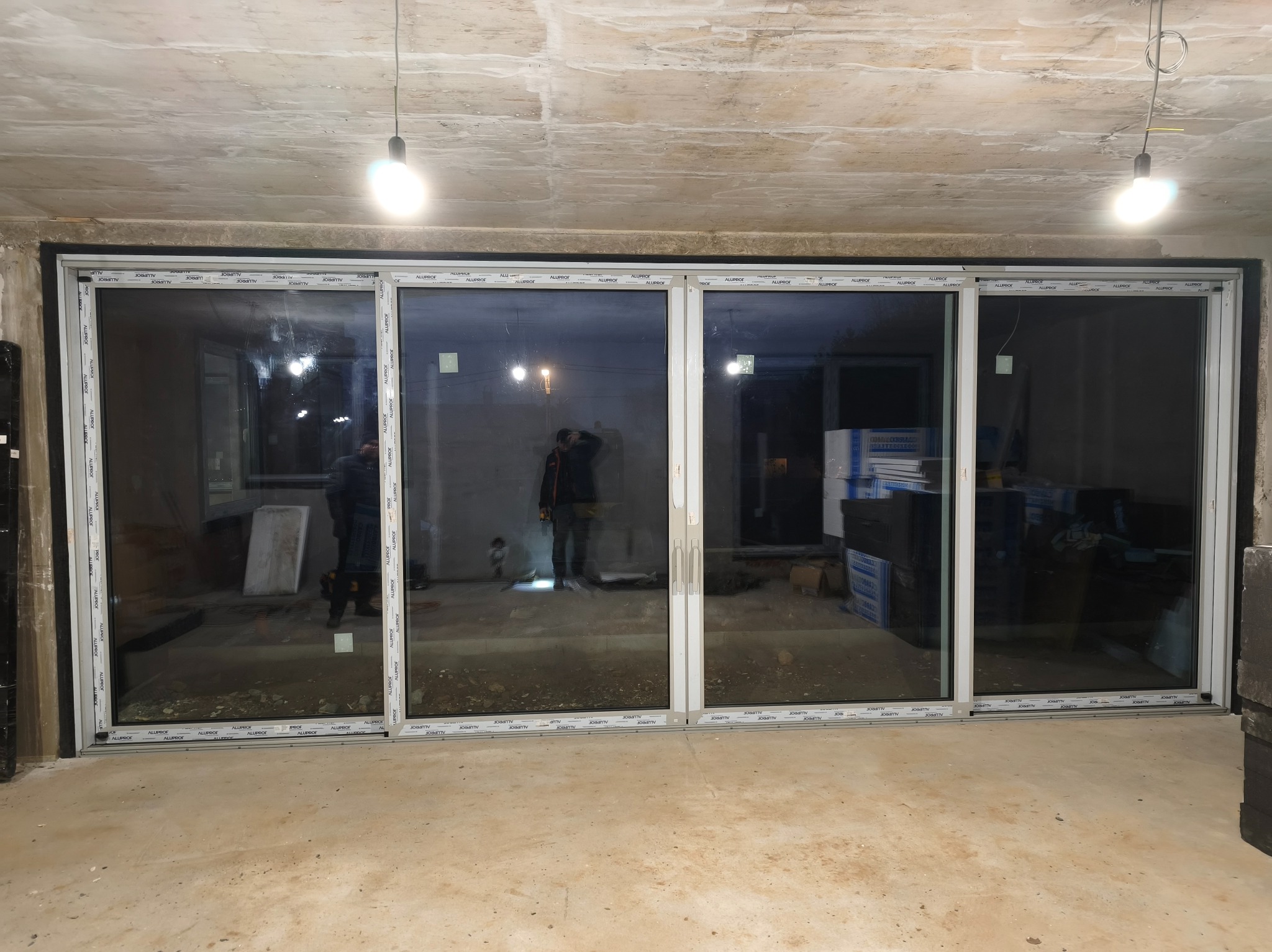 Czteroskrzydłowe drzwi przesuwne z białymi profilami Aluplast, zamontowane w surowym wnętrzu z betonowym sufitem i podłogą. Widoczne odbicia osób w szybach.