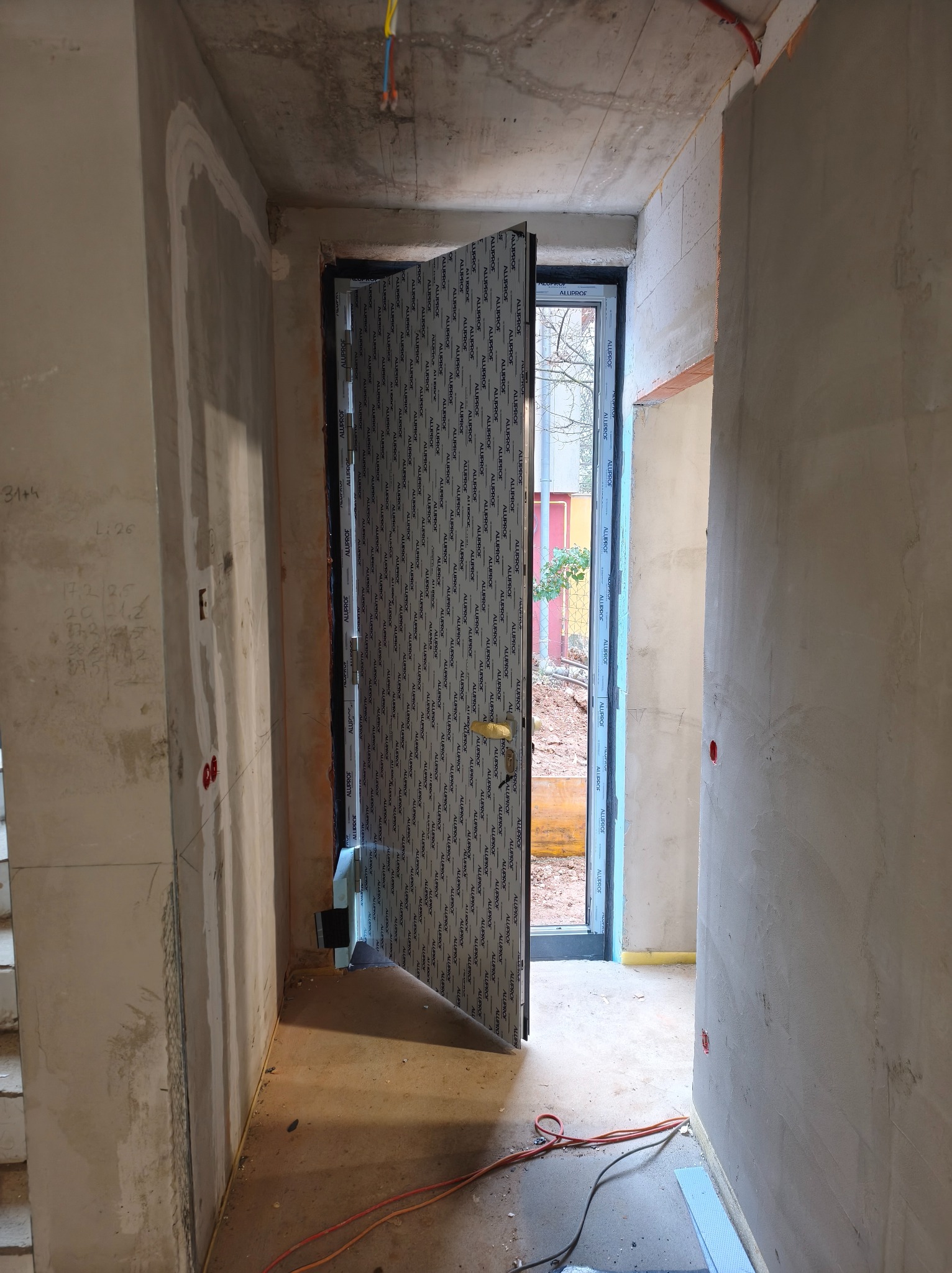 Otwarta, szara, zewnętrzna aluminiowa drzwi z szybą, z widocznym logo producenta, w surowym wnętrzu budynku w trakcie wykończenia. Widok na zewnątrz.