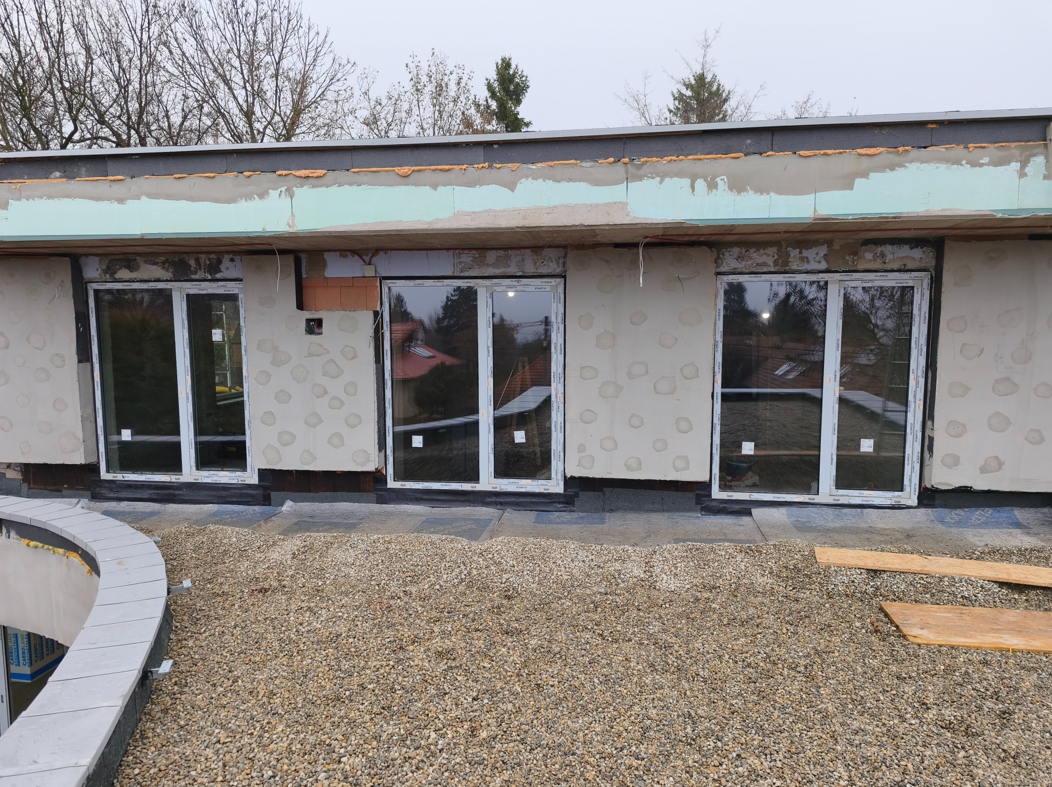 Montaż okien PCV na etapie wykończenia elewacji budynku. Widoczne ocieplenie, surowa ściana z naklejonym styropianem i trzy okna z białymi ramami. Na dole żwir.