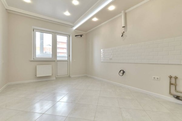Jasne wnętrze po remoncie: białe płytki na ścianie, kremowe ściany, okno balkonowe, grzejnik i jasna podłoga z płytek. Widoczne instalacje elektryczne i hydrauliczne.