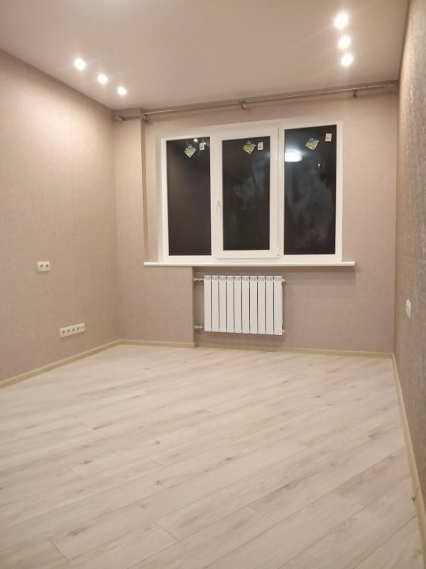 Jasne, puste wnętrze pokoju po remoncie: panele podłogowe, beżowe ściany, białe okno z grzejnikiem. Widoczne gniazdka elektryczne i oświetlenie sufitowe.