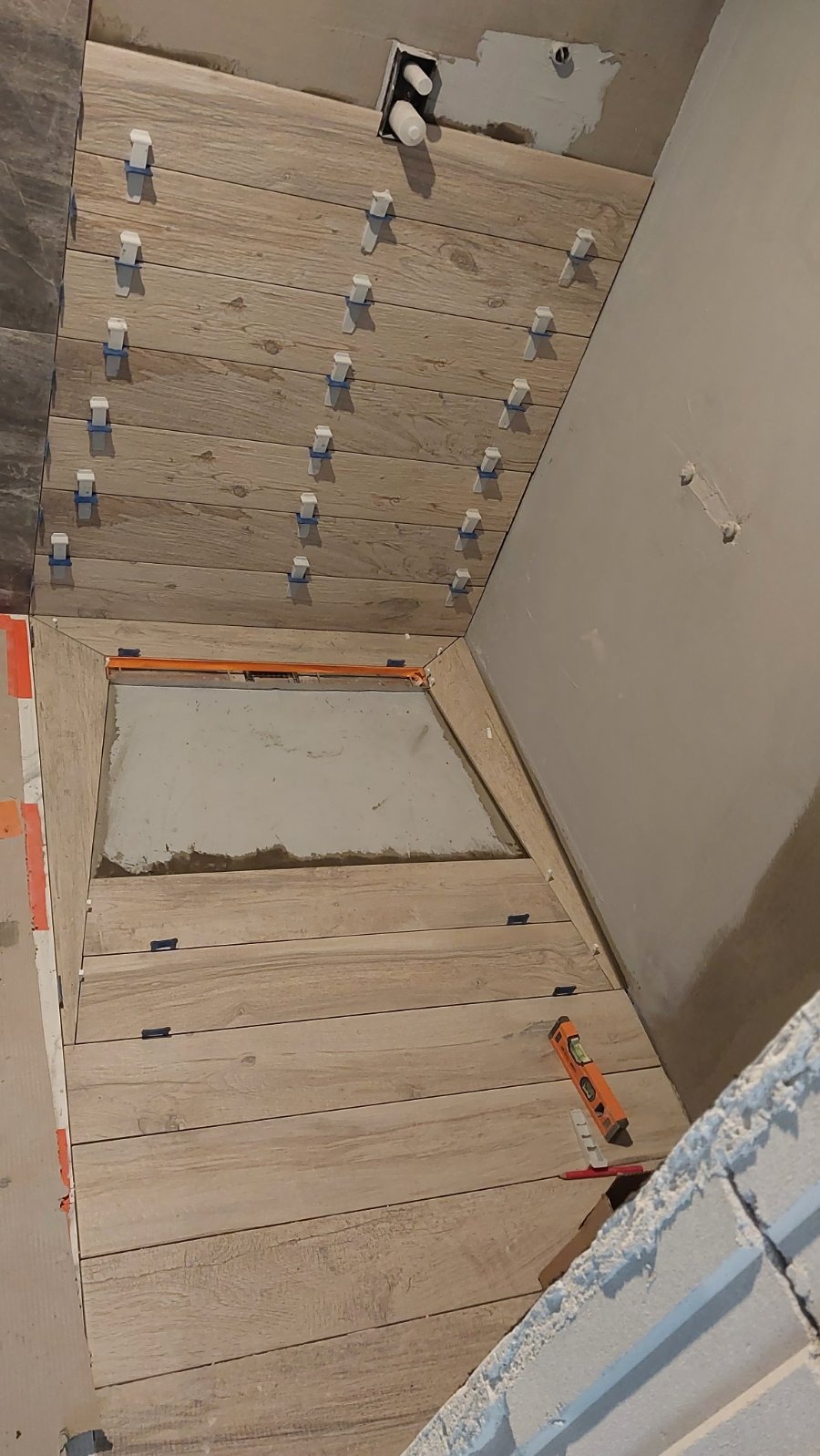 Układanie płytek imitujących drewno w łazience, z użyciem poziomicy i klipsów do wyrównywania. Widoczny fragment brodzika i instalacja wodna w ścianie.